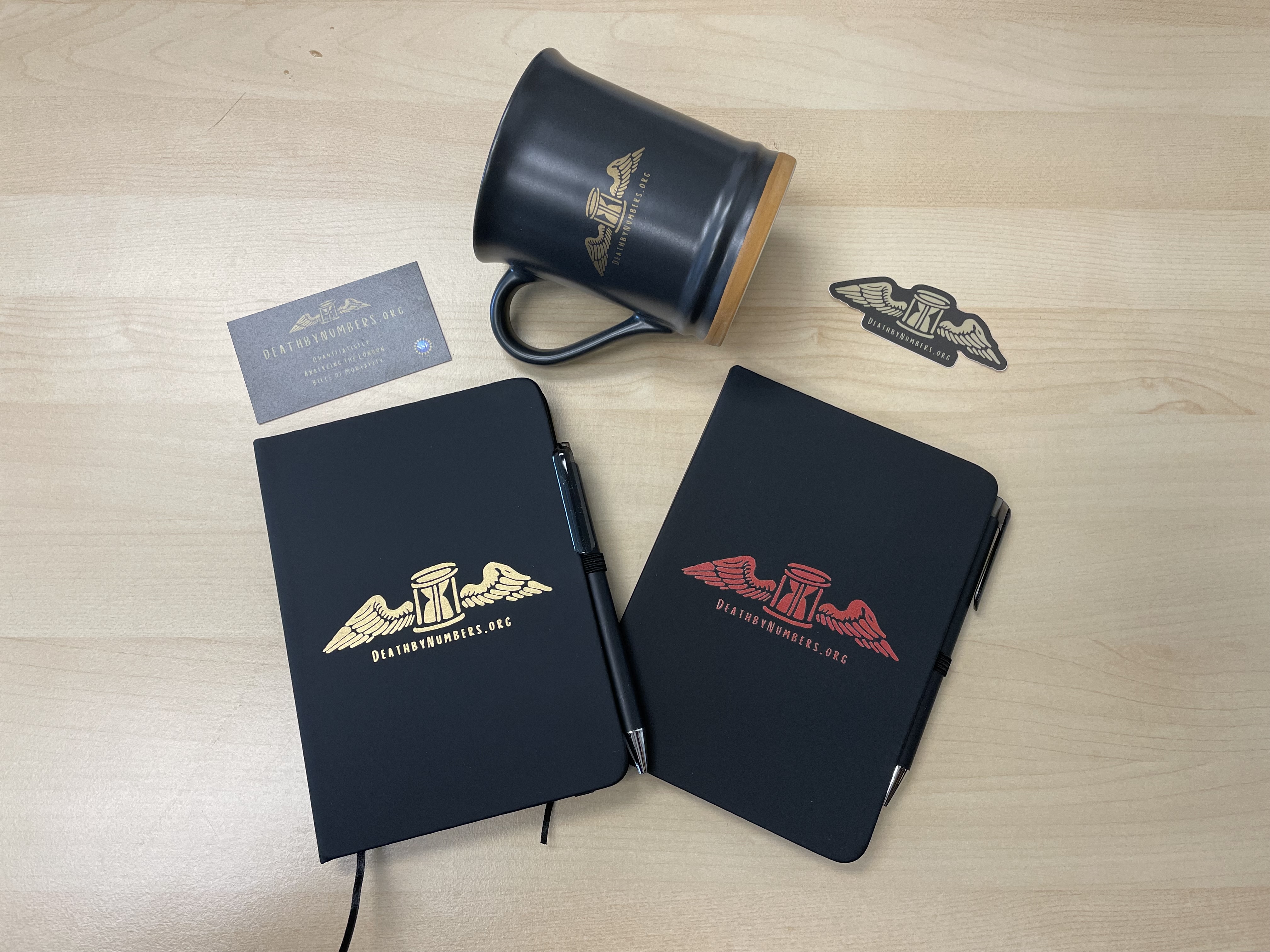 black business card with gold logo and writing, black mug with gold logo, black sticker with gold logo, and two black notebooks with gold and red logos; the logo is a winged hourglass with the url deathbynumbers.org below it